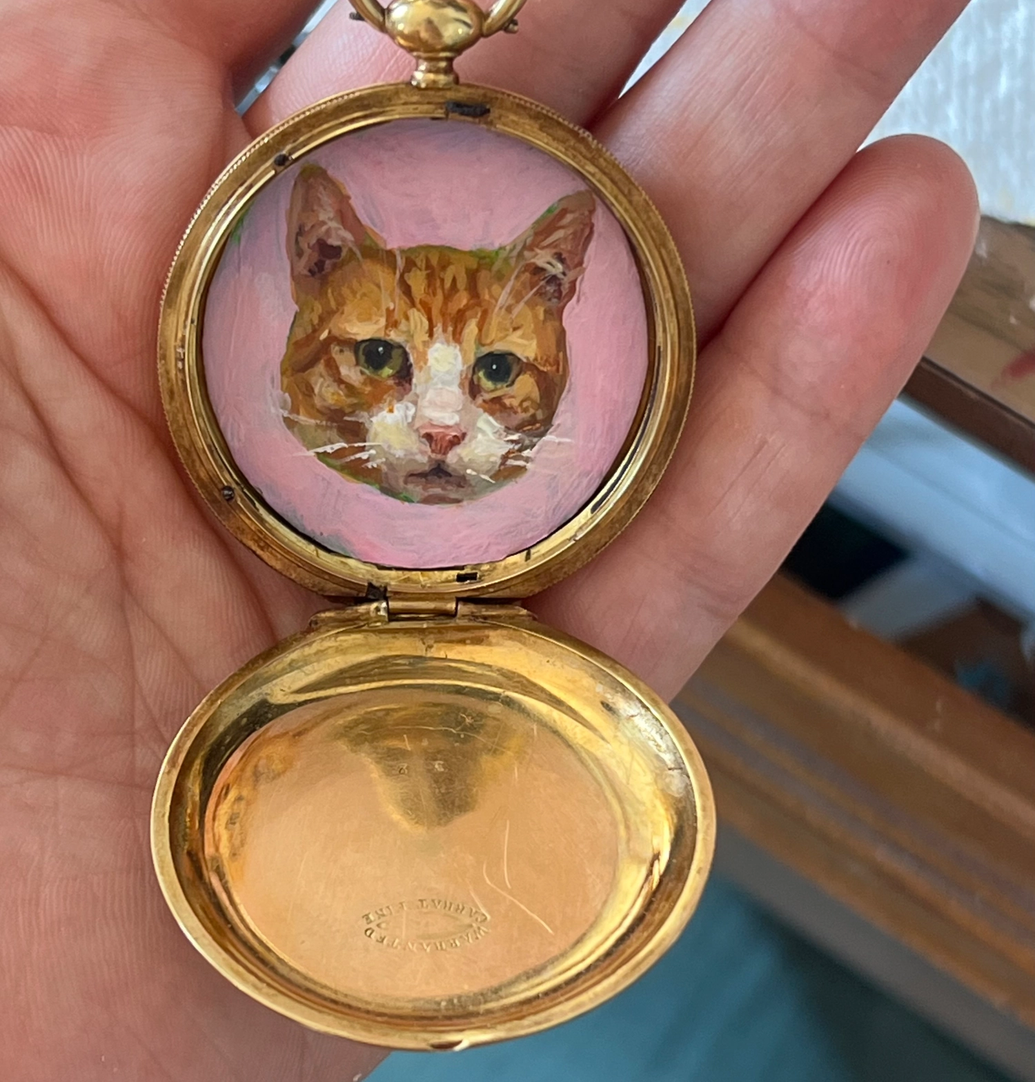 A painting of an orange cat. The painting is set in an empty gold pocket watch.